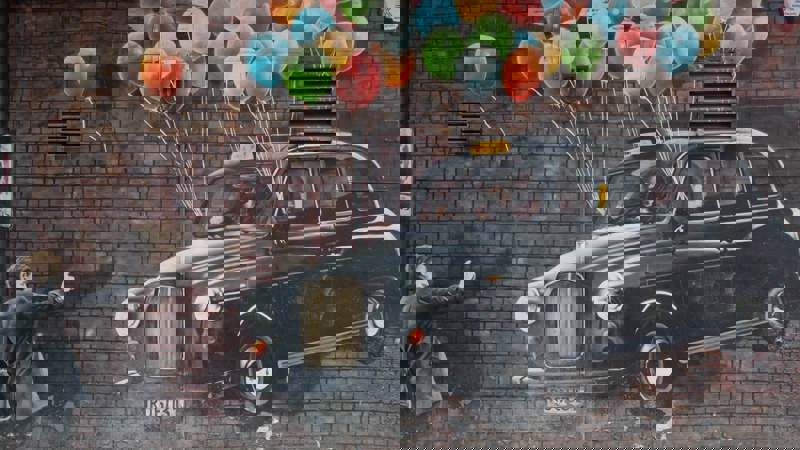 A mural on a brick wall showing a man in a black taxi being lifted into the air by colorful balloons.