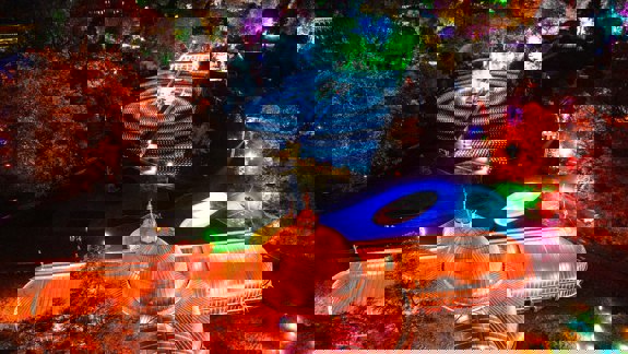 An aerial night view of the illuminated Kibble Palace and surrounding Botanic Gardens, featuring bright orange and pink lights, and a swirling blue light display on the grass.