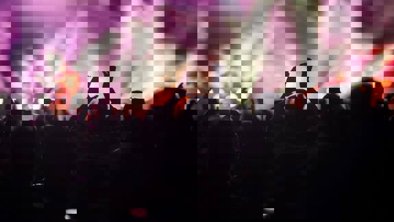 A cheering concert audience with hands raised, silhouetted against purple and white stage lights.