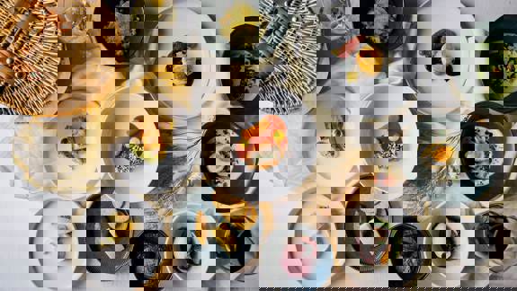 A variety of gourmet dishes are displayed on a white tablecloth, including a cheeseboard, appetizers, and several small plates. A picnic basket and glasses of wine are also visible.