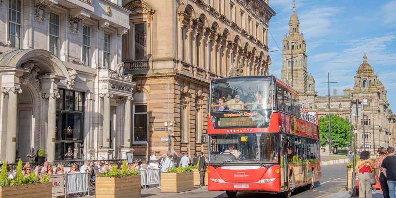 A red open-top tour bus drives past historic buildings in Glasgow on a sunny day, with people dining outdoors and walking nearby.