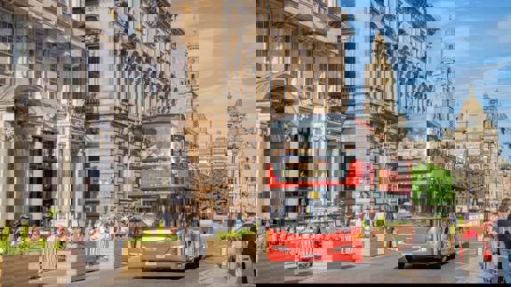 A red open-top tour bus drives past historic buildings in Glasgow on a sunny day, with people dining outdoors and walking nearby.