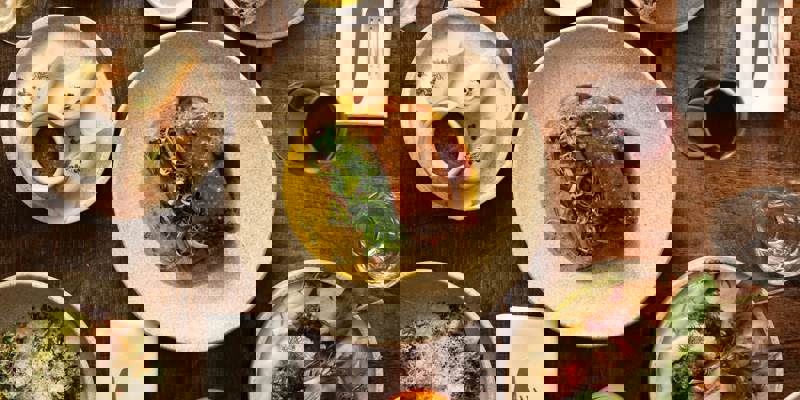 An overhead shot of several plates of gourmet food and drinks arranged on a rustic wooden table. One plate features a piece of meat with a green sauce.