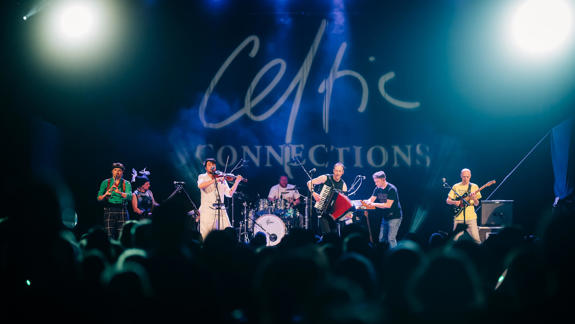 A Celtic folk band performing on stage with various instruments under bright stage lights, with "Celtic Connections" displayed in the background.