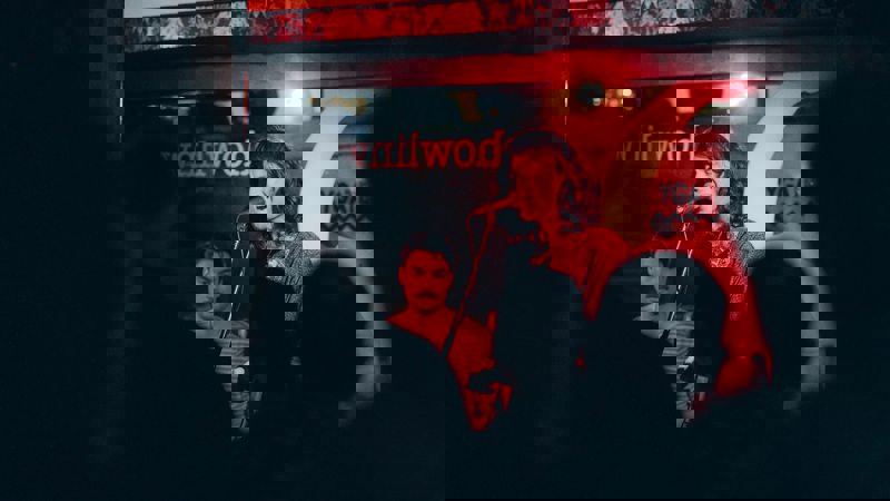 A singer with long, dark hair performs on a stage lit by red light, with a bass player behind him.