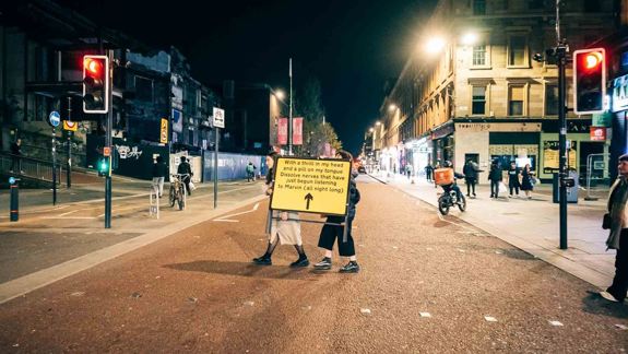 People walk at night across Sauchiehall Street holding a large yellow sign with text. Illuminated buildings and traffic lights create an urban ambience.