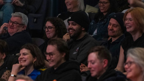 A diverse audience smiling and laughing while watching a screening at the Glasgow Film Festival (GFF), seated in dark lighting.