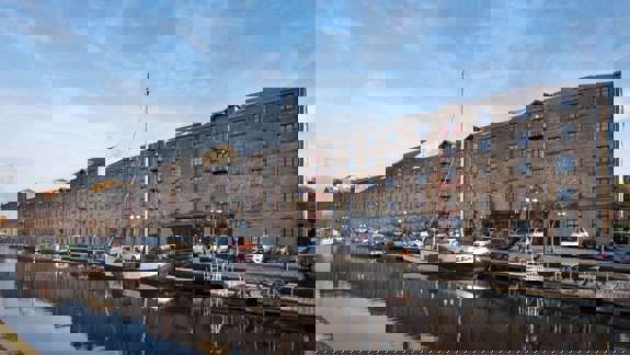 A long, multi-story building with many windows sits alongside a canal with a boat docked. Cars are parked along the water's edge.