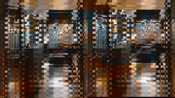 A sophisticated, low-lit bar with dark wood finishes, leather armchairs, candlelit tables, and a moody ambiance designed for quiet evening drinks.