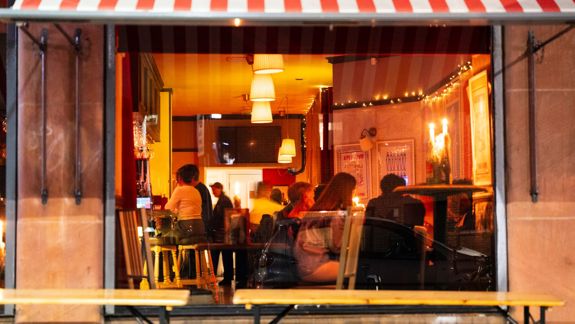 The warm, inviting interior of Bar Vini is visible through the window, showing patrons enjoying drinks and conversations under a red and white awning.
