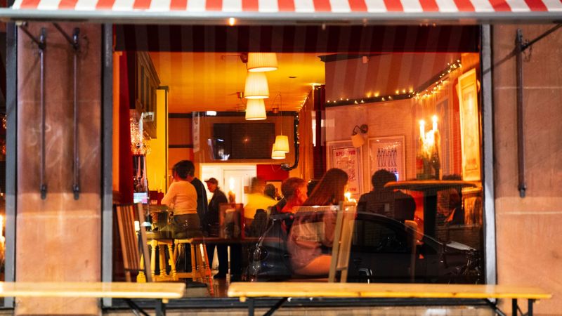 The warm, inviting interior of Bar Vini is visible through the window, showing patrons enjoying drinks and conversations under a red and white awning.