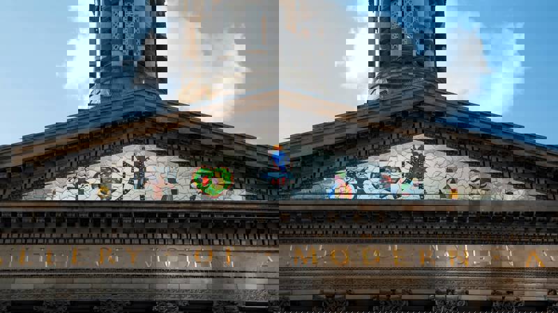 A colourful stained-glass mosaic depicting abstract figures in the tympanum of a building labelled "GALLERY OF MODERN ART."