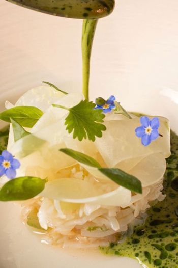 A close-up of a white, intricately plated dish with edible blue flowers, being finished with a pour of vibrant green oil sauce.
