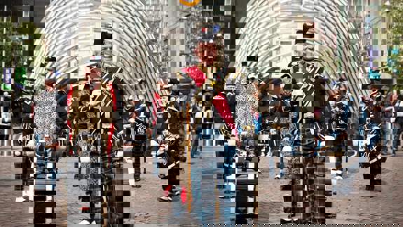 A Scottish pipe band in full dress tartan, marching down a cobbled street with large drums.