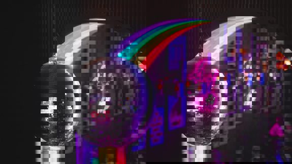 A disco ball hangs in the foreground with a rainbow archway and people in a bar in the background.