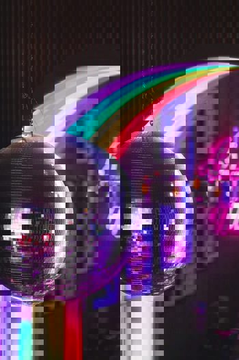 A disco ball hangs in the foreground with a rainbow archway and people in a bar in the background.