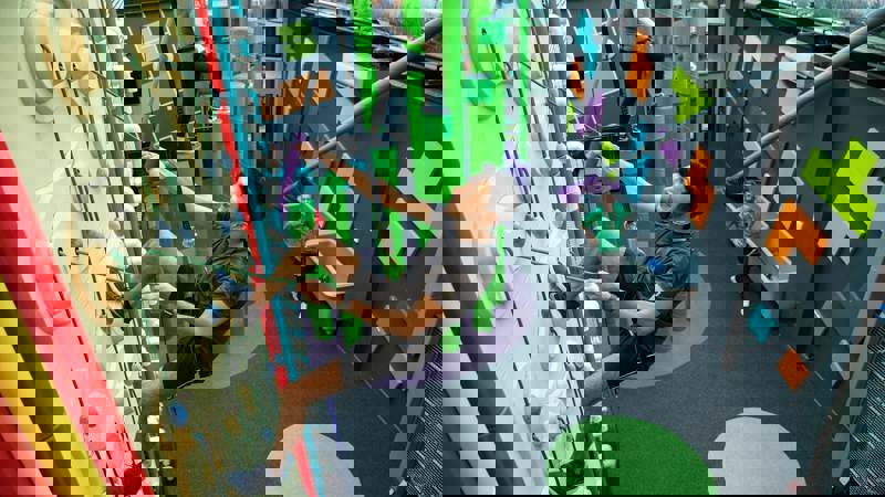 An athletic man is indoor rock climbing with a harness and rope. Another person is climbing on green poles in the background, and another person is on the ground.