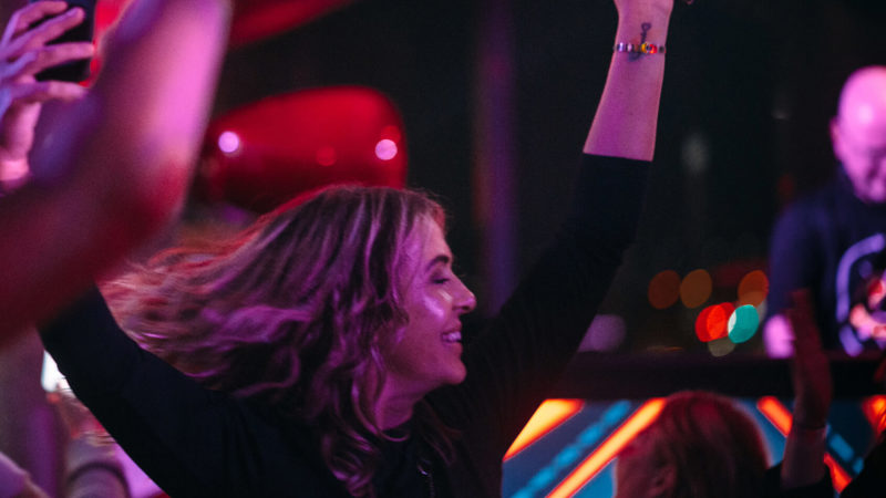 Person smiling broadly and raising their hands in the air as they dance, surrounded by an energetic crowd at a vibrant indoor party with colourful lights.