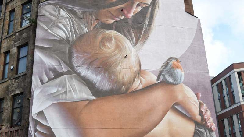 A large mural on the side of a building depicts a woman holding a baby close, with a small robin perched on the baby's arm.
