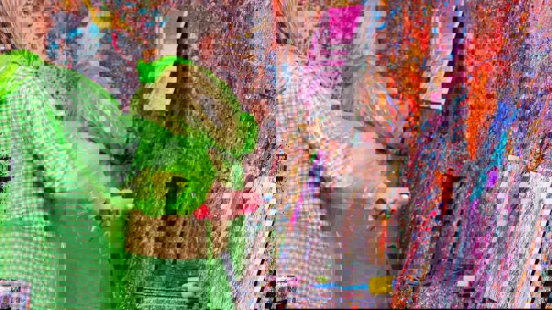 Two individuals wearing protective ponchos are in a room covered in colorful paint splatters, working on their own canvases at a splatter paint studio.