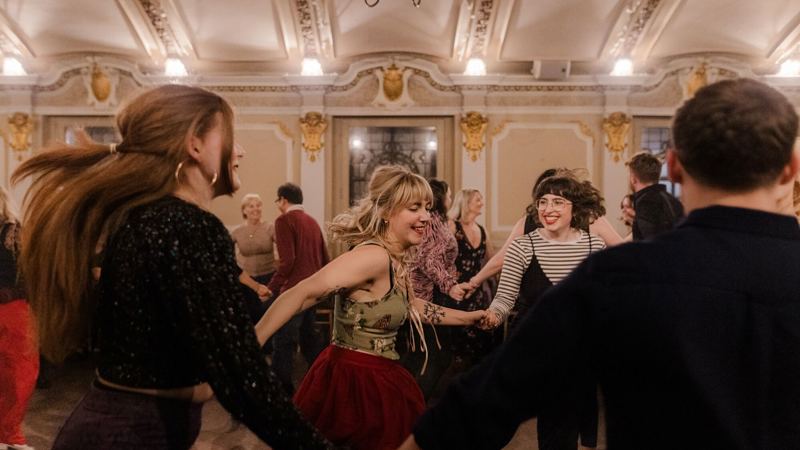 A joyful Scottish ceilidh dance in a grand, ornate hall. People hold hands, spinning and laughing in a lively atmosphere under chandeliers.