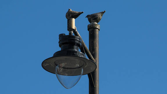 Two bird sculptures perched on a lamppost, silhouetted against a clear blue sky.