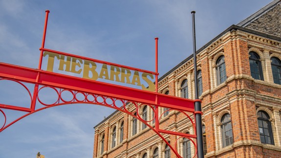 A red gantry sign reading 'The Barras' against a blue sky and an ornate stone building.