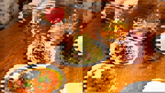 A close-up of a wooden table laden with various plates of food and drinks. The dishes include a salad with shredded vegetables, a bright orange dish, and polenta fries, with wine glasses and tumblers.