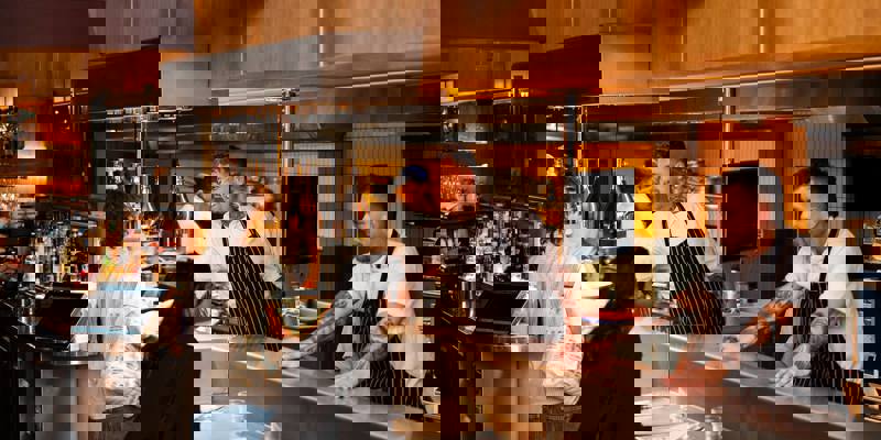 An indoor shot of a busy restaurant counter with chefs and customers interacting. Two chefs are behind the counter, smiling, while a customer stands in front of it.