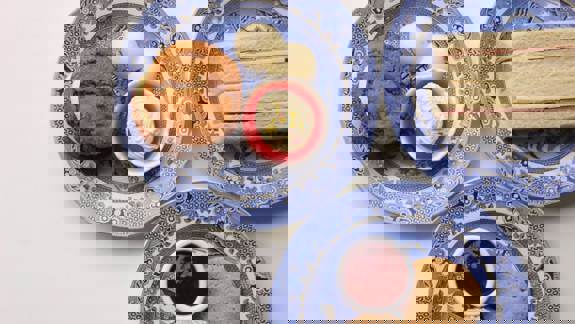 Plates of sandwiches, scones with cream and jam, and assorted desserts on patterned plates on an off-white table setting.