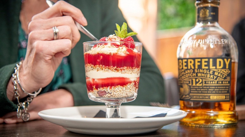 A person eating a dessert of raspberries, cream, and oats next to a bottle of Aberfeldy whisky.