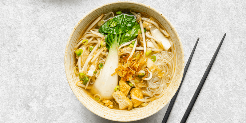 Bowl of clear noodle soup with bok choy, bean sprouts, green onions, and tofu, placed on gray surface with black chopsticks