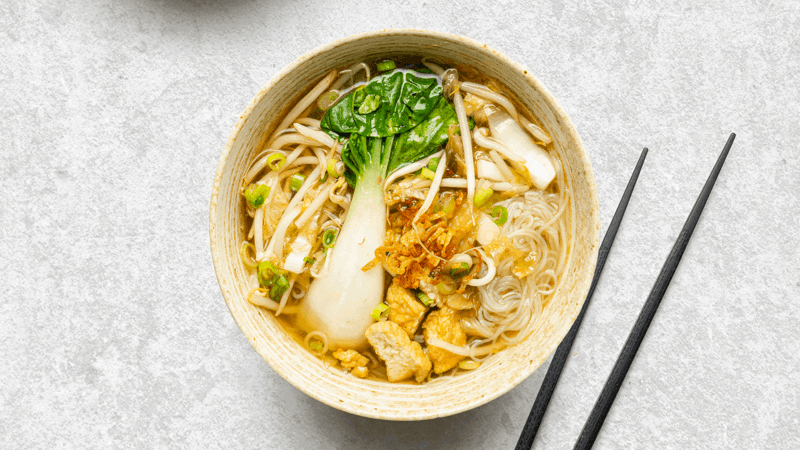 Bowl of clear noodle soup with bok choy, bean sprouts, green onions, and tofu, placed on gray surface with black chopsticks
