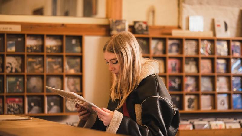 A person with long blond hair looks at a record album, with rows of albums visible on shelves in the background.