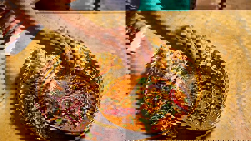 A hand squeezing lemon onto a rich curry dish served in a small brass karahi, alongside flaky parathas and a side salad, all on a gold platter.