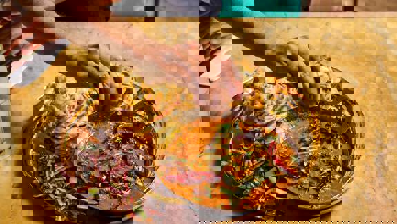 A hand squeezing lemon onto a rich curry dish served in a small brass karahi, alongside flaky parathas and a side salad, all on a gold platter.