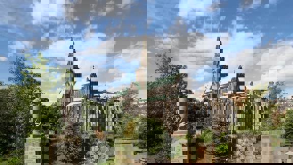 The historic Glasgow Cathedral, a Gothic stone building, stands against a blue sky with white clouds. The foreground includes gravestones and trees.
