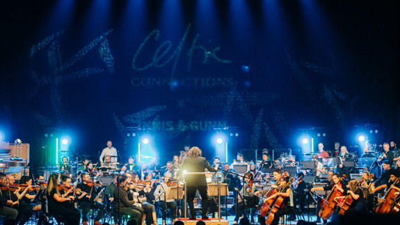 A vibrant orchestra performs on stage under blue lights at Celtic Connections, with a conductor leading, and the event logo visible in the background.