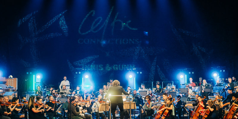 A vibrant orchestra performs on stage under blue lights at Celtic Connections, with a conductor leading, and the event logo visible in the background.
