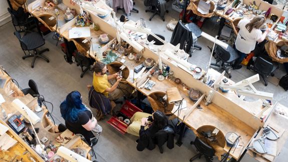 Aerial view of a busy workshop with cluttered wooden desks in rows. People are engaged in crafting, surrounded by tools and materials, conveying creativity.