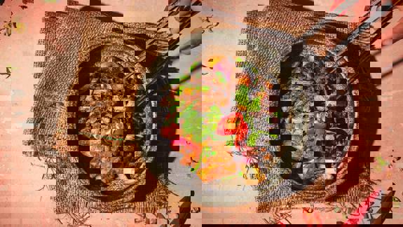 A close-up, overhead shot of a vibrant, spicy stir-fry or curry in a dark bowl, being picked up with chopsticks.