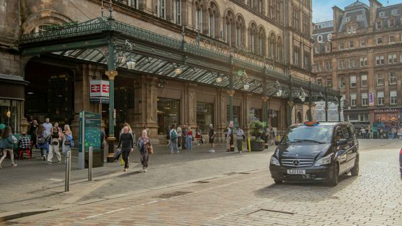 People walking outside a large historic train station with a taxi driving on a cobblestone street.