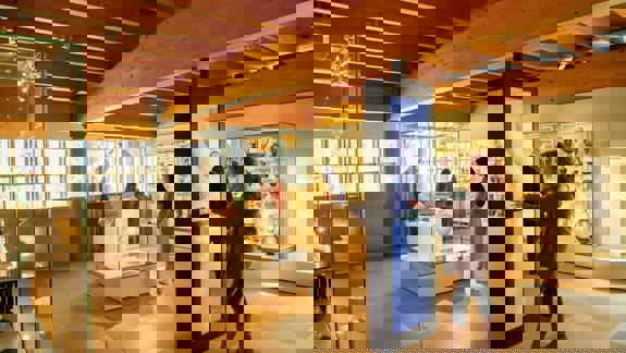  Visitors interacting with an upright digital screen and viewing ceramic and ancient artefacts displayed in glass cases under a wooden-beamed ceiling at The Burrell Collection.