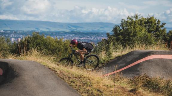 A mountain biker in a black t-shirt and red helmet rides on a dirt track with a city and hills in the background.