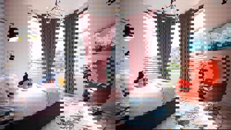 Bright, modern hotel bedroom with a large bed, an orange Egg Chair, pink curtains in a bay window, and a stylish gold and black light fixture.