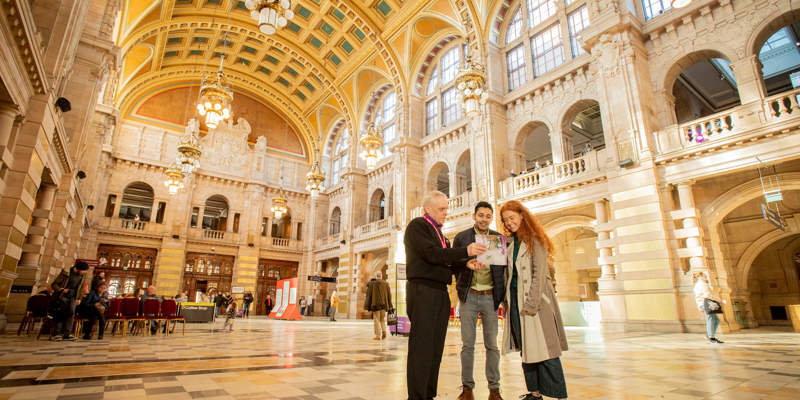 A museum staff member shows a map to two visitors in a vast, ornate hall with high vaulted ceilings, golden chandeliers, and arched balconies.