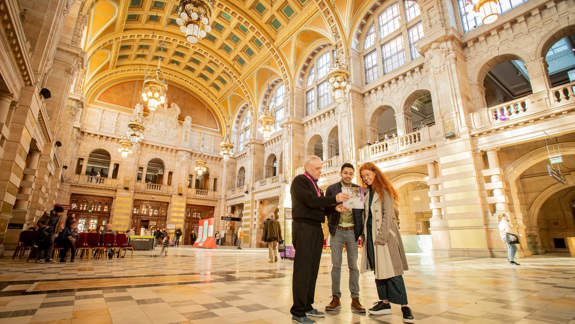 A museum staff member shows a map to two visitors in a vast, ornate hall with high vaulted ceilings, golden chandeliers, and arched balconies.