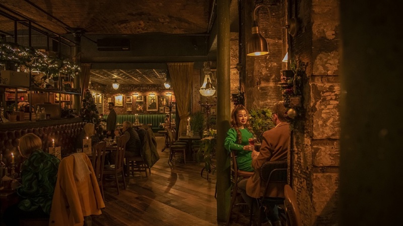 A couple talks in a dark, cosy pub with exposed brick, wood floors, and warm, low lighting from candles and lamps.