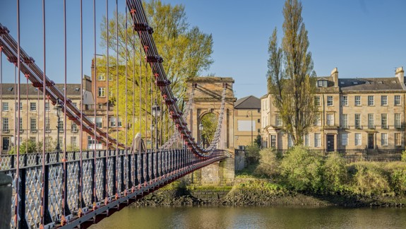 A red suspension bridge across a river. Victorian era sandstone buildings and trees line the riverbank.