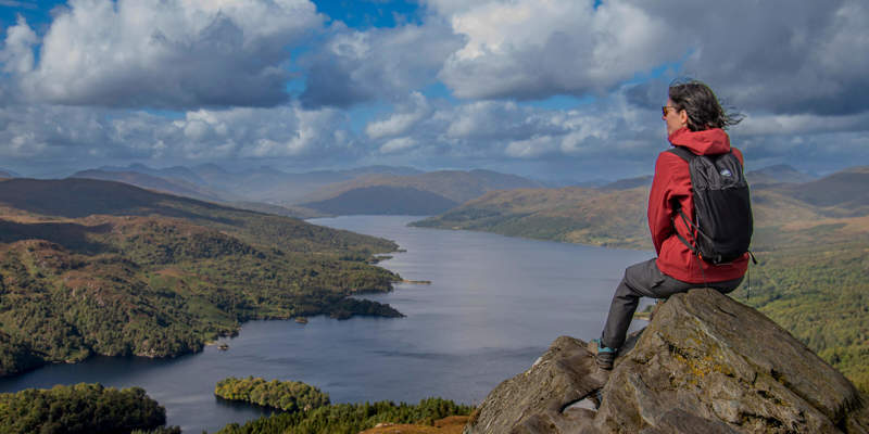 Hiker on Ben A'an summit overlooks Loch Katrine and the stunning Trossachs National Park on a sunny day.